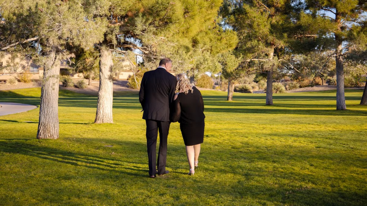 A couple walking arm-in-arm in a sunny park, surrounded by trees and grass.
