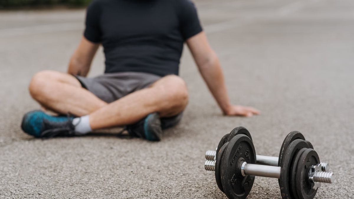 Crop anonymous male athlete in sports clothes sitting with crossed legs on asphalt roadway near dumbbells after training