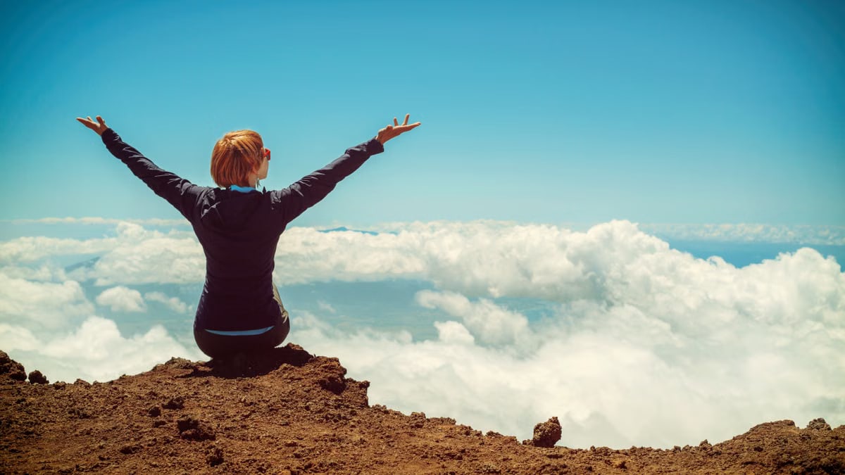A woman enjoys a scenic view atop a cliff in Kula, Hawaii, surrounded by clouds and blue sky.