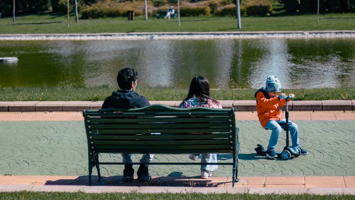 A family relaxes on a bench by the pond while a child plays on a scooter.